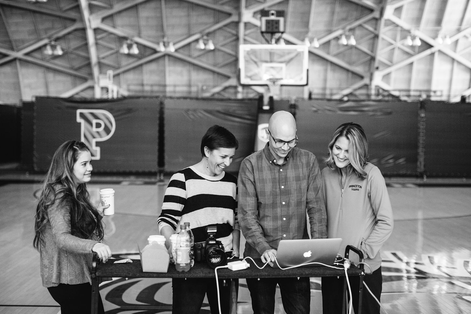 Four people collaborate around a laptop and equipment on a table inside a gymnasium in black and white.
