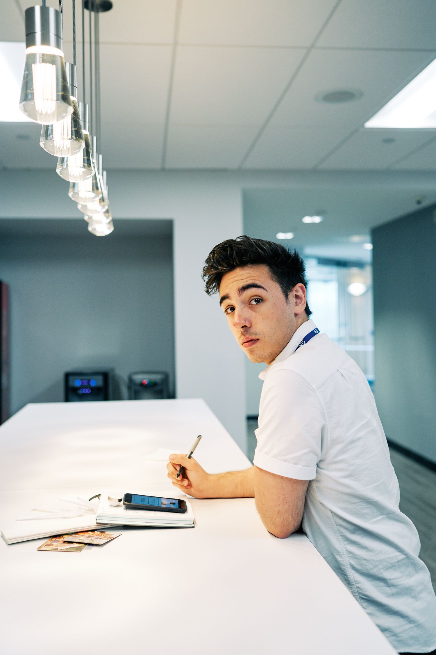 Young man in white shirt writing notes at a modern office desk with pendant lights overhead.