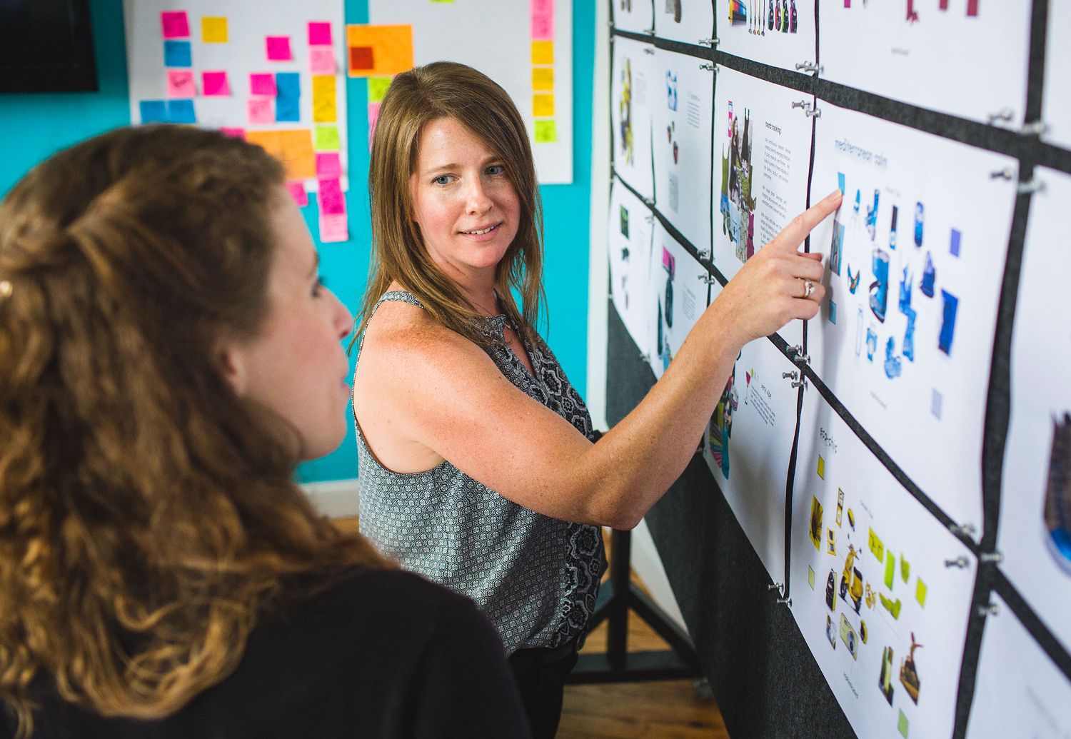 Two women collaborating and reviewing design documents pinned to a large office planning board.