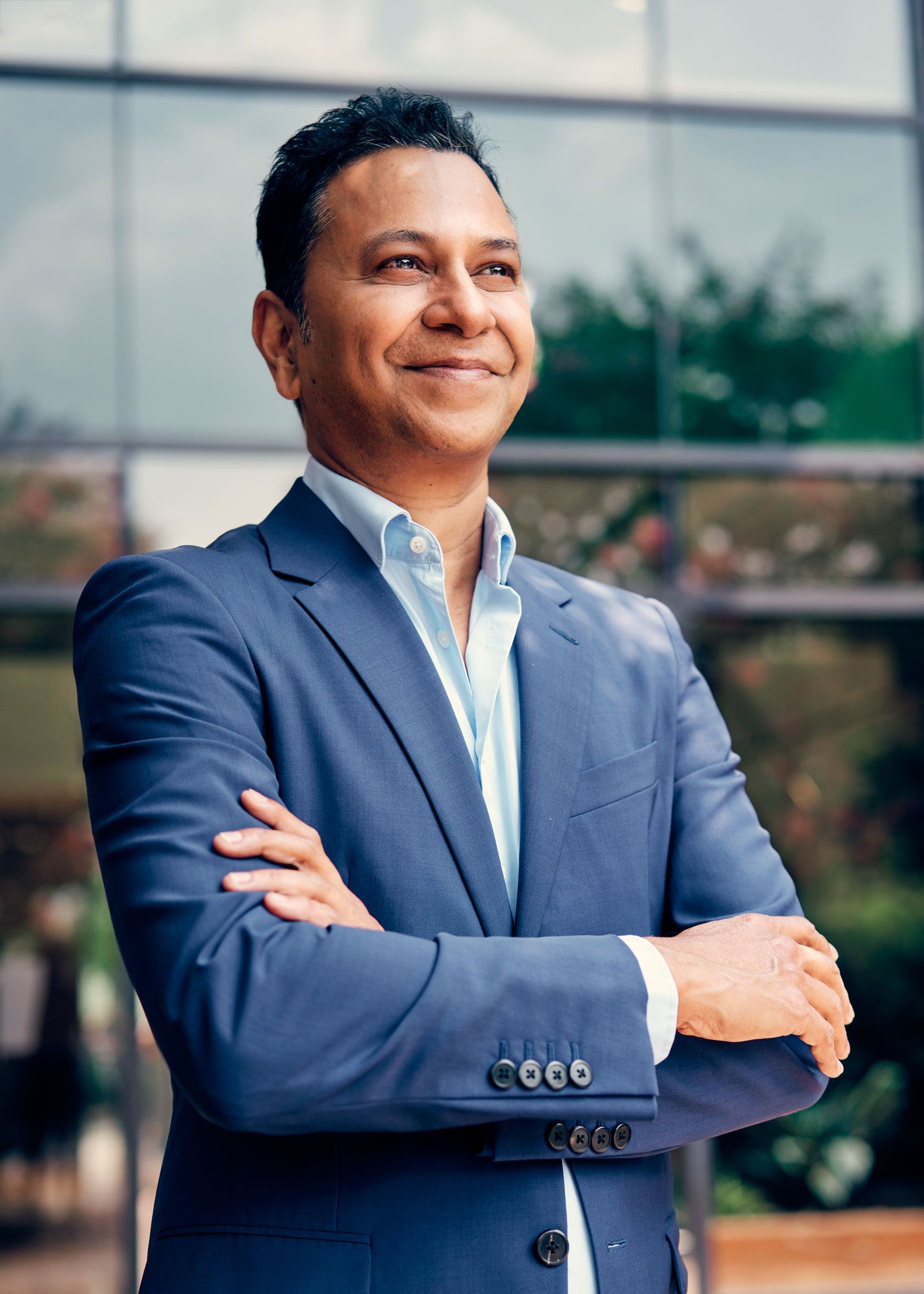 Confident businessman in blue suit stands with arms crossed smiling outdoors near glass building.