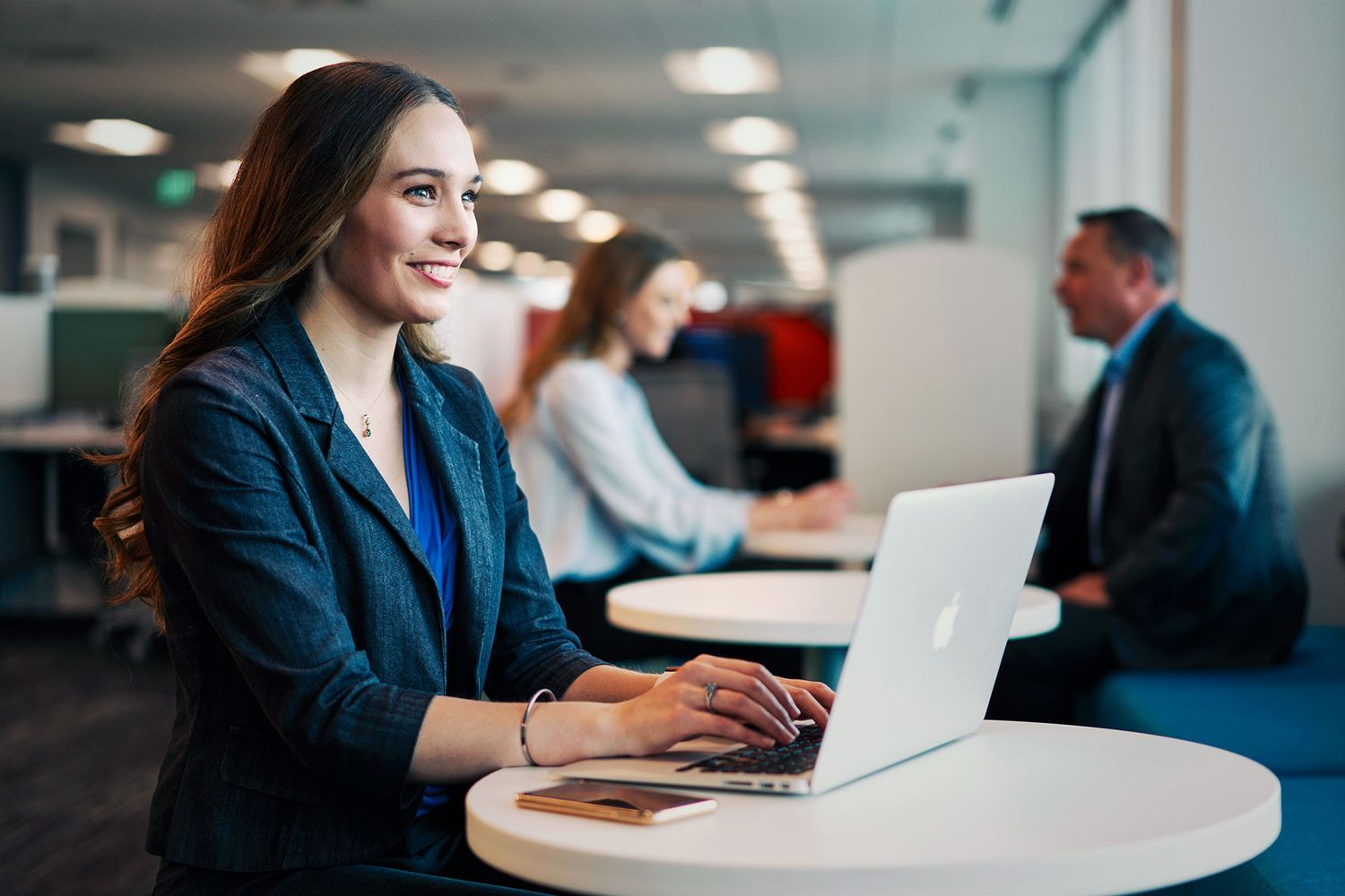 Young businesswoman smiling while working on laptop in modern open-plan office with colleagues in background.