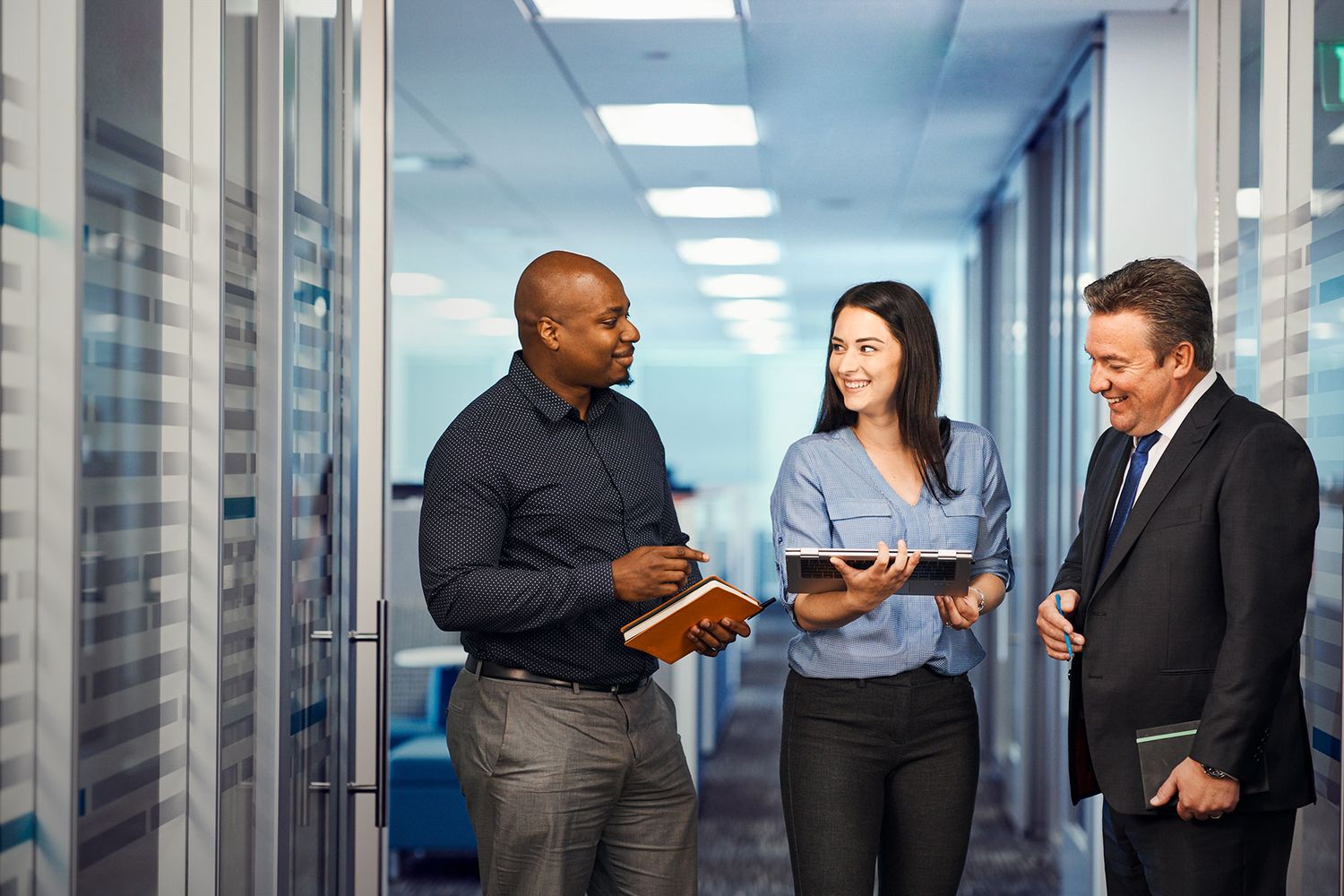 Three business professionals discussing work in a modern office hallway, with two holding tablets.