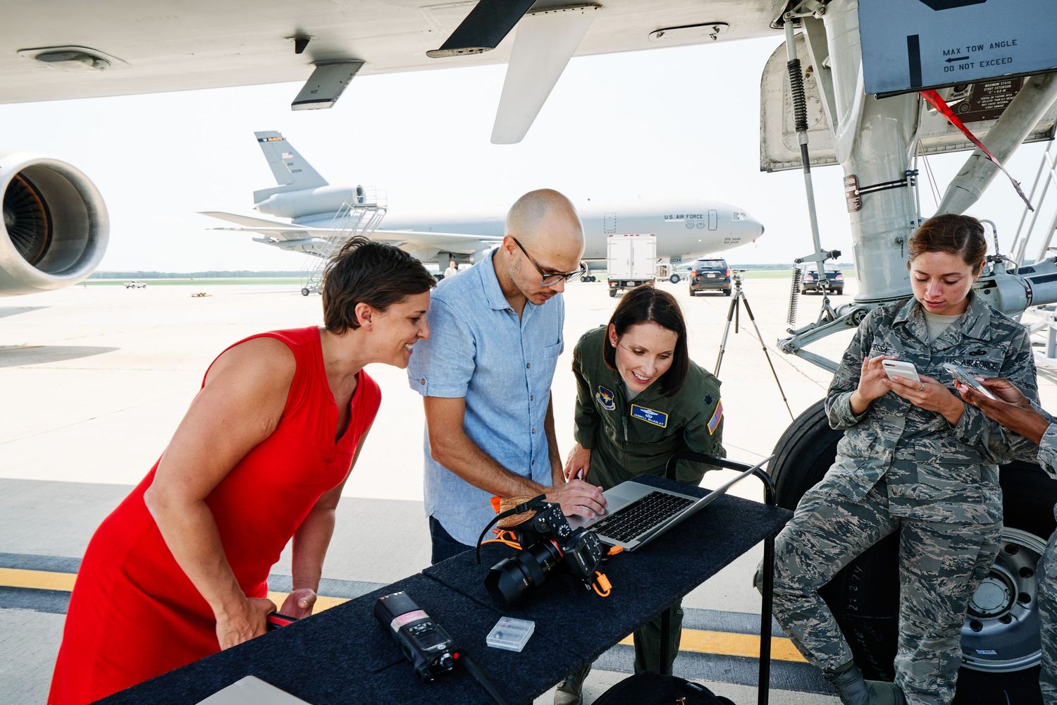 Four people reviewing laptop data on tarmac beside military aircraft and drone equipment.
