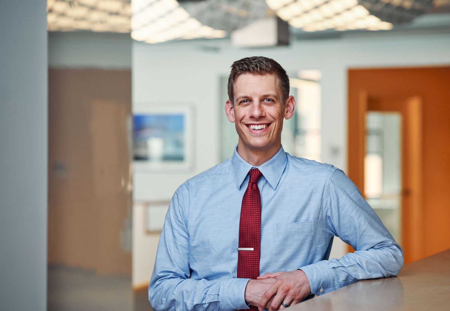Confident young businessman in blue shirt and red tie smiling while leaning on a counter in a bright office.