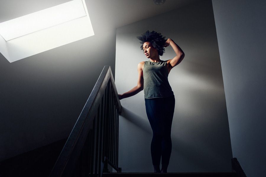 A woman in athletic wear stands on stairs under a skylight, striking a confident pose in a dramatic, moody interior.
