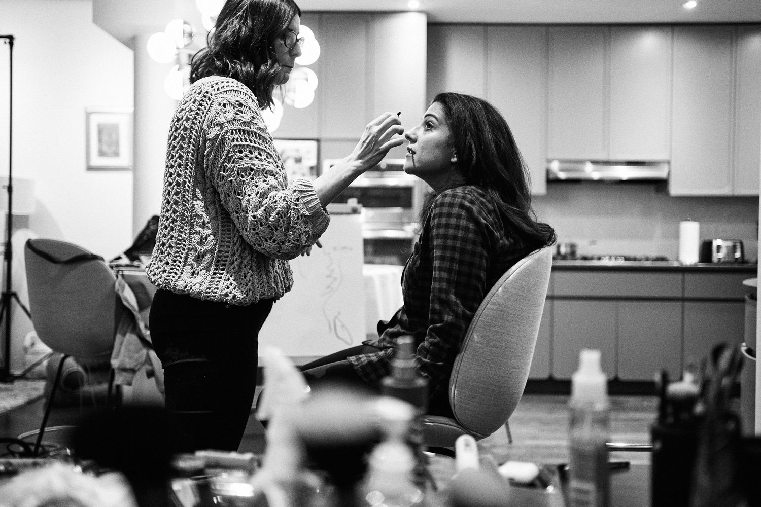 Black and white photo of a makeup artist applying makeup to a woman seated in a vanity chair in a modern kitchen.