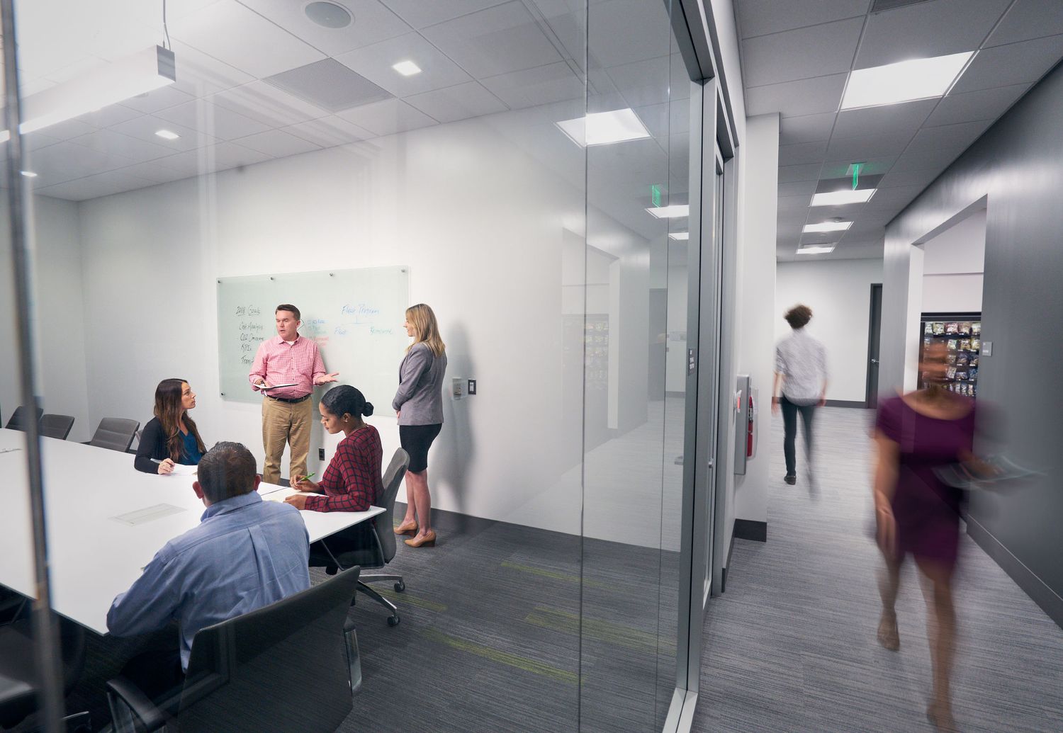 Team collaborates in modern glass-walled conference room while colleague walks through bright office hallway.