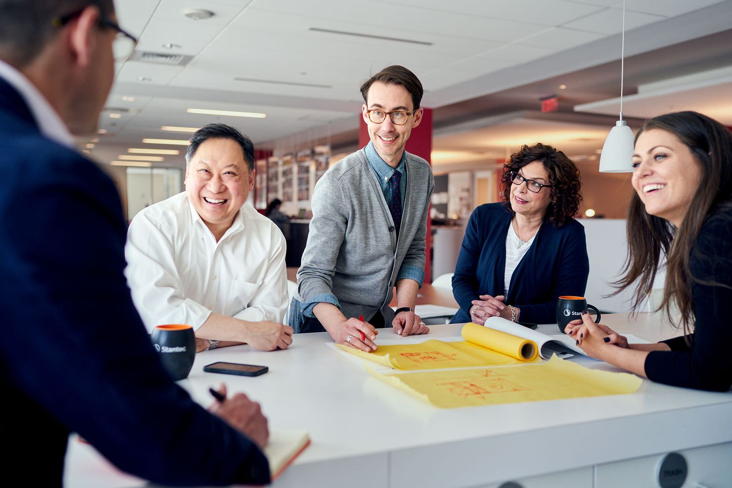 A diverse group of five business professionals collaborating over architectural blueprints at a bright modern office table.