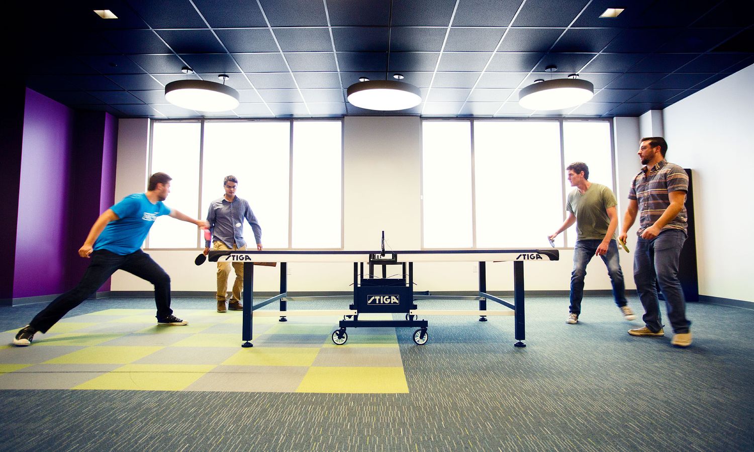 Four colleagues enjoy a competitive ping pong game in a colorful modern office break room.