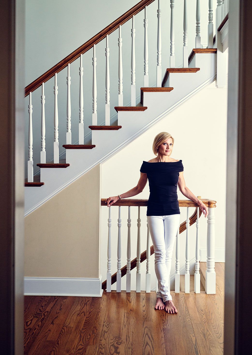 Blonde woman in black top and white jeans poses by elegant staircase in bright home.