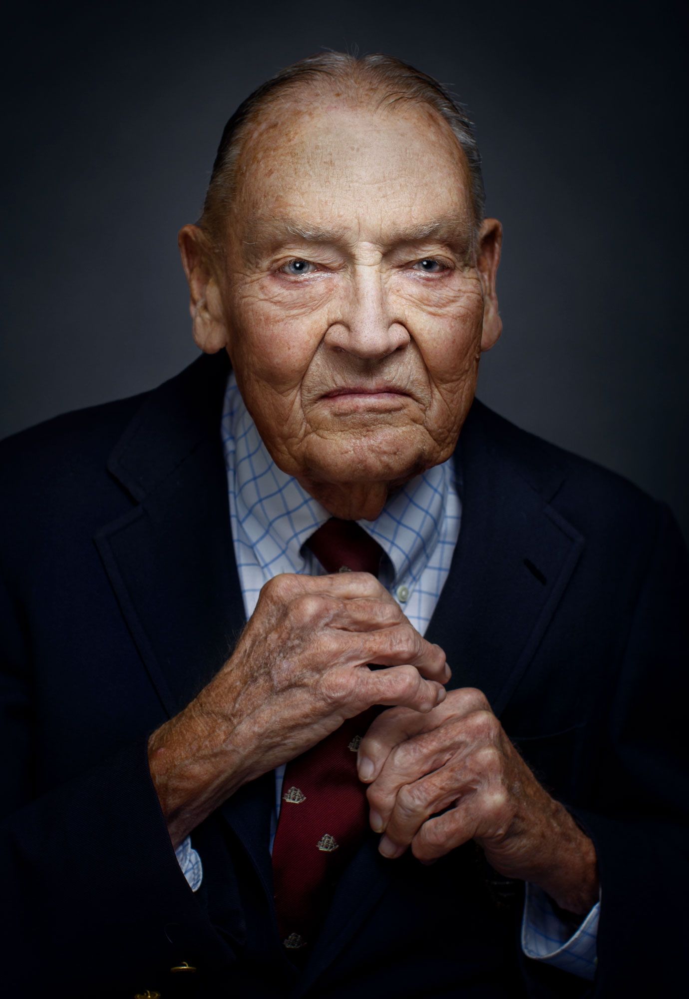Close-up portrait of elderly man in navy suit with clasped hands against dark background.