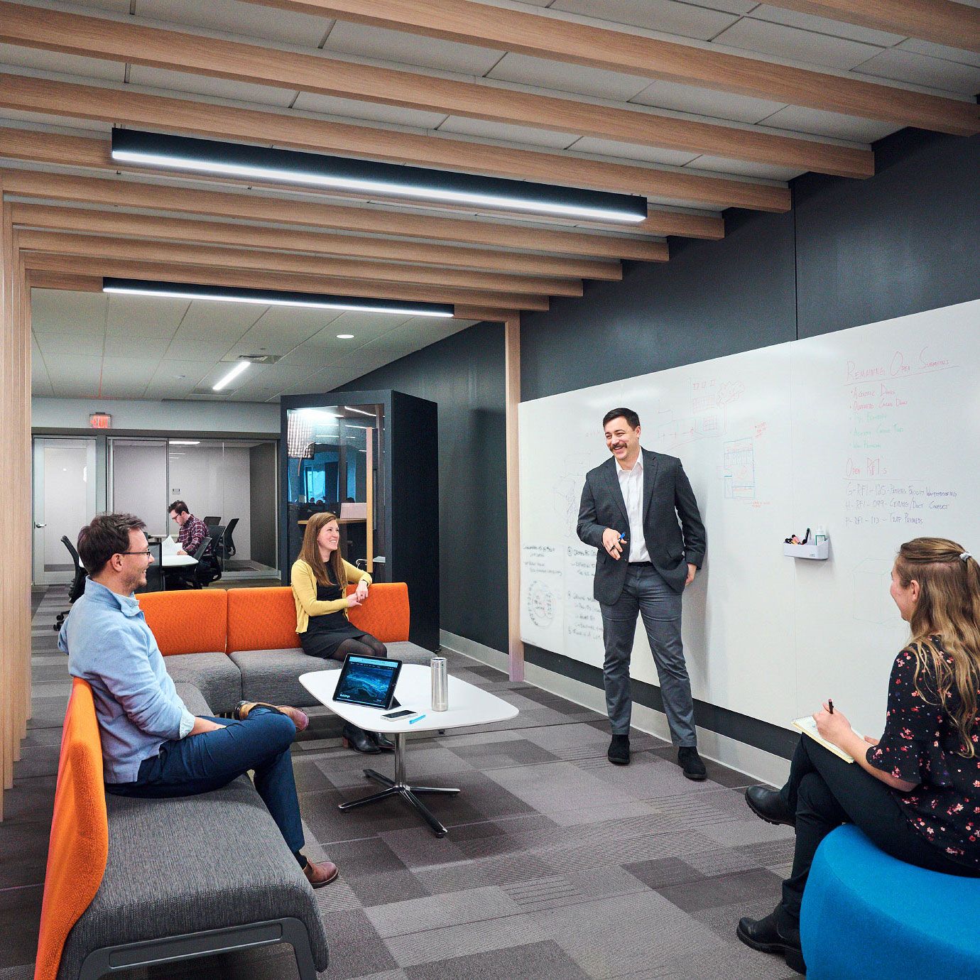 Modern office collaboration space with whiteboard presentation, colorful seating, and team members engaged in discussion.
