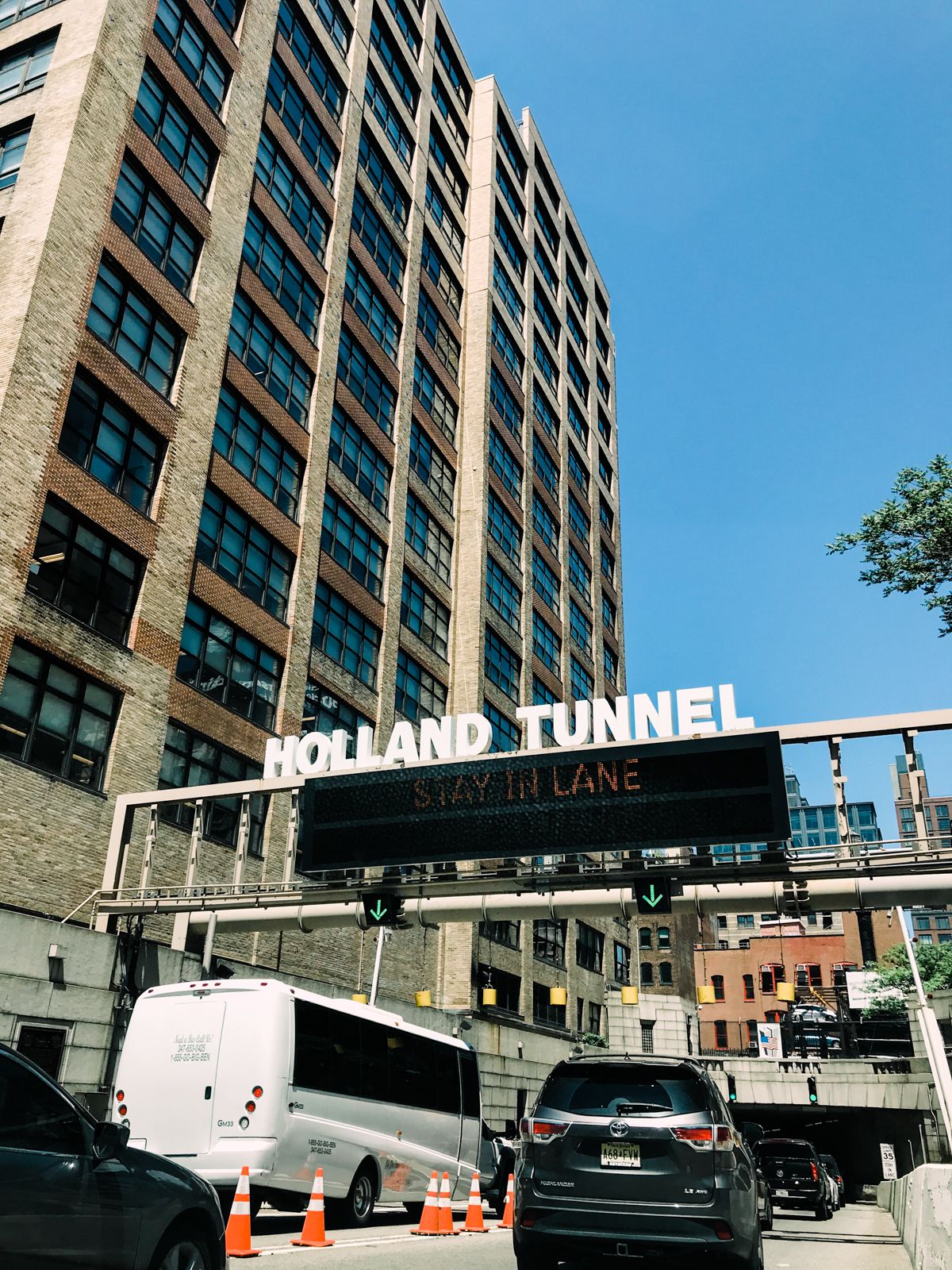 Cars and buses queue at the Holland Tunnel entrance in NYC, with orange traffic cones and a large overhead sign on a sunny day.