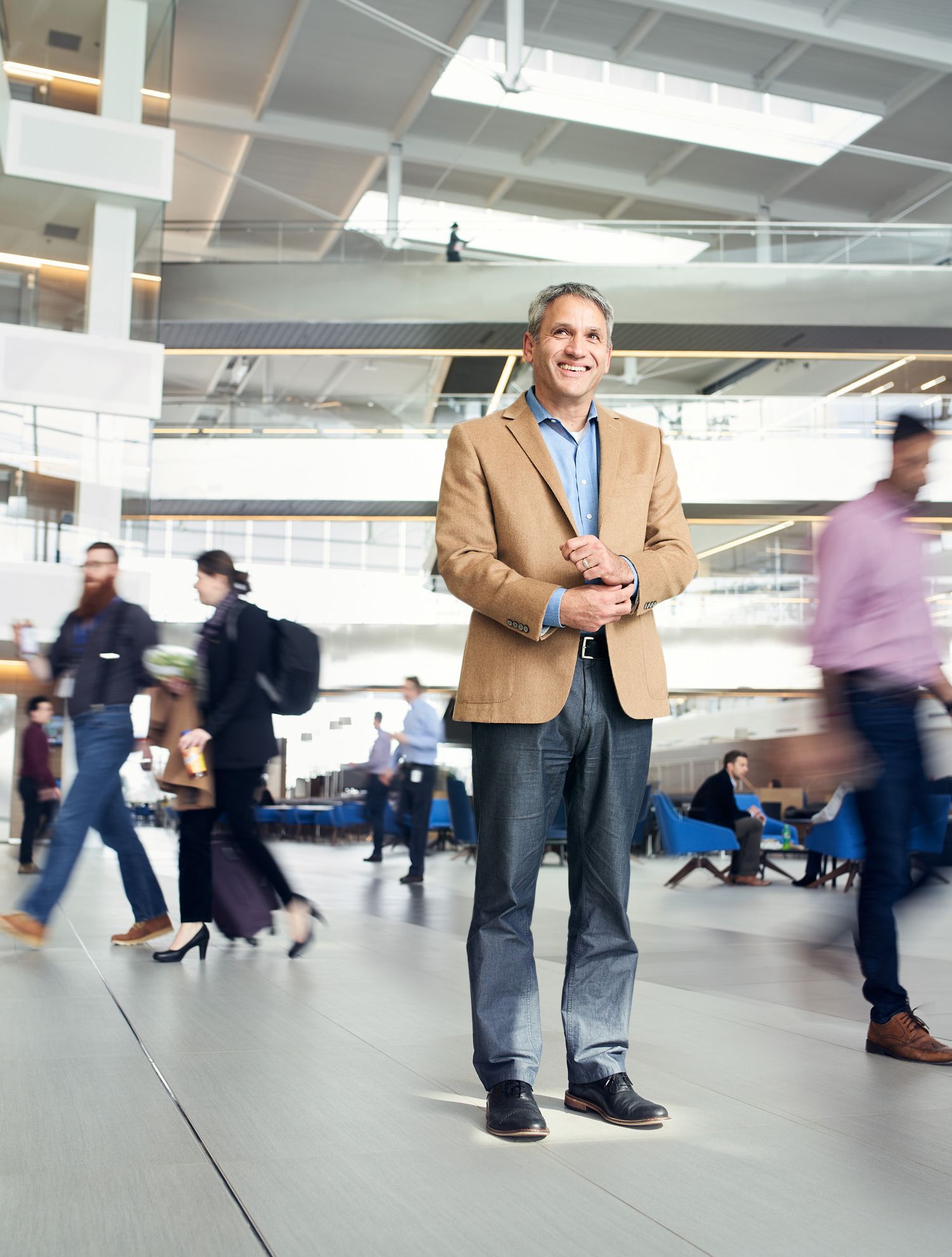 Smiling man in tan blazer and jeans stands confidently in busy modern office atrium with blurred people walking around him.