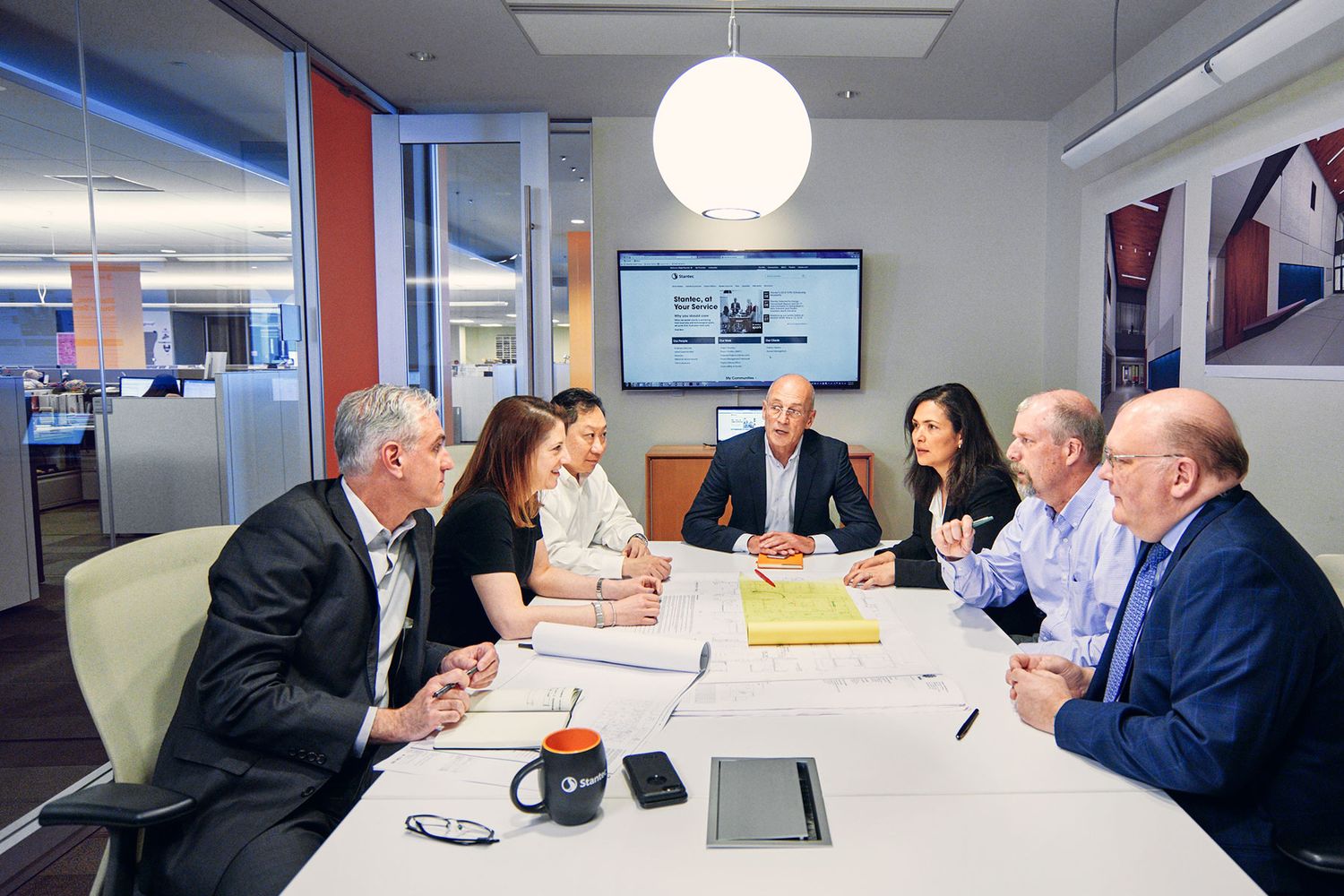 Seven business executives engaged in a formal meeting around a white conference table in a modern office.