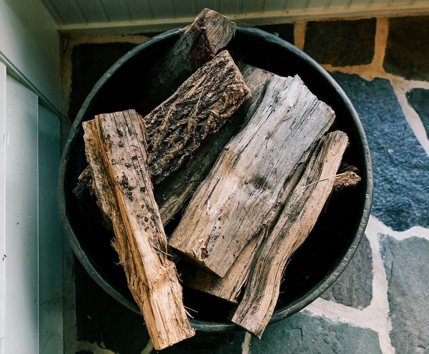 Rustic firewood logs stacked in a dark metal bucket near a stone wall.