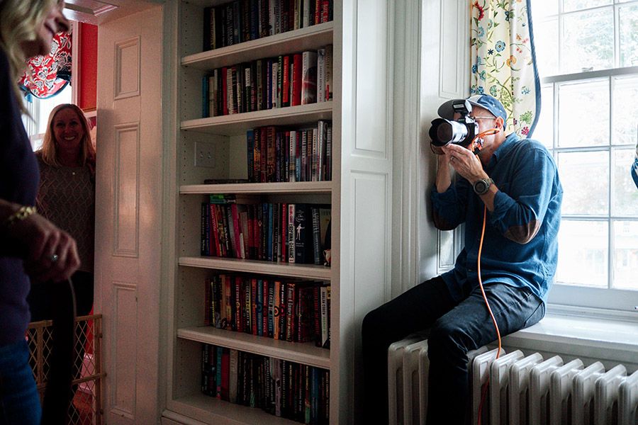A photographer crouches on a windowsill with a camera, capturing shots beside a built-in bookshelf filled with books.