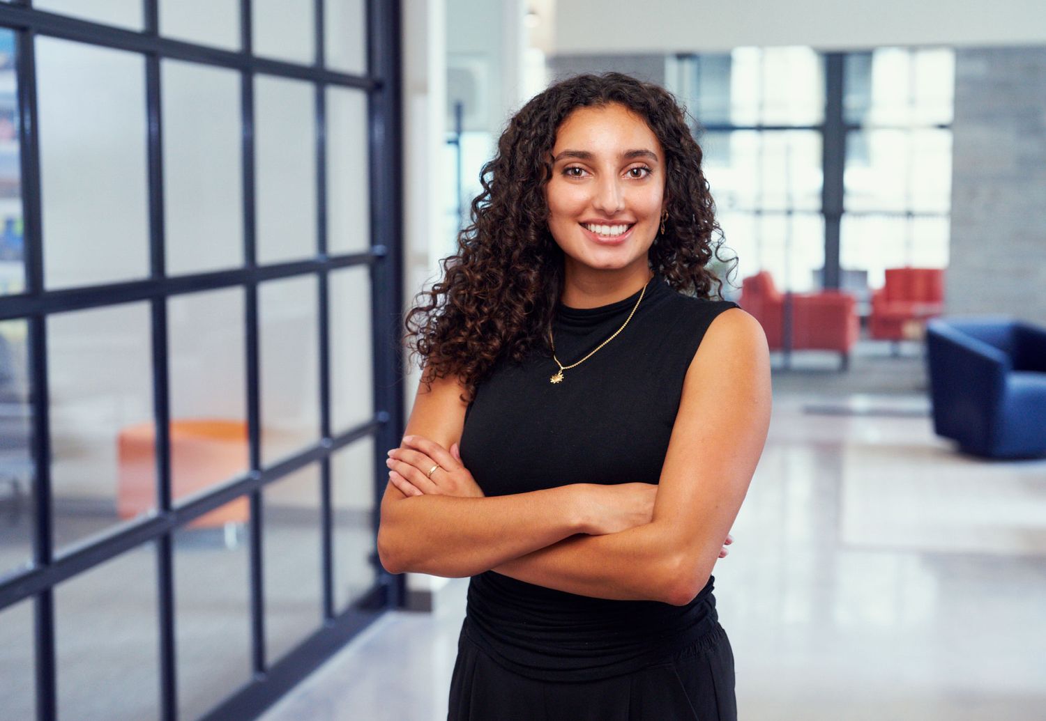 Young professional woman with curly hair stands confidently with arms crossed in a modern office lobby.