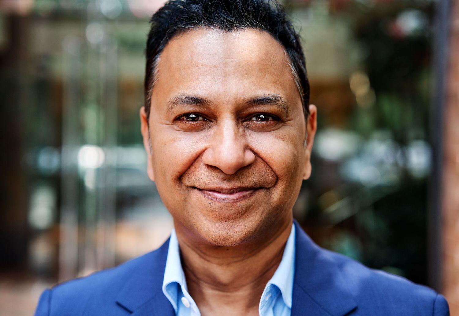 Confident smiling man in blue blazer poses outdoors in urban setting with greenery background.