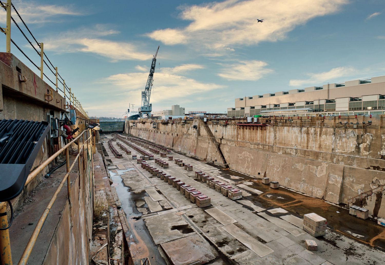 Empty dry dock with concrete walls, crane, and industrial buildings under partly cloudy sky.