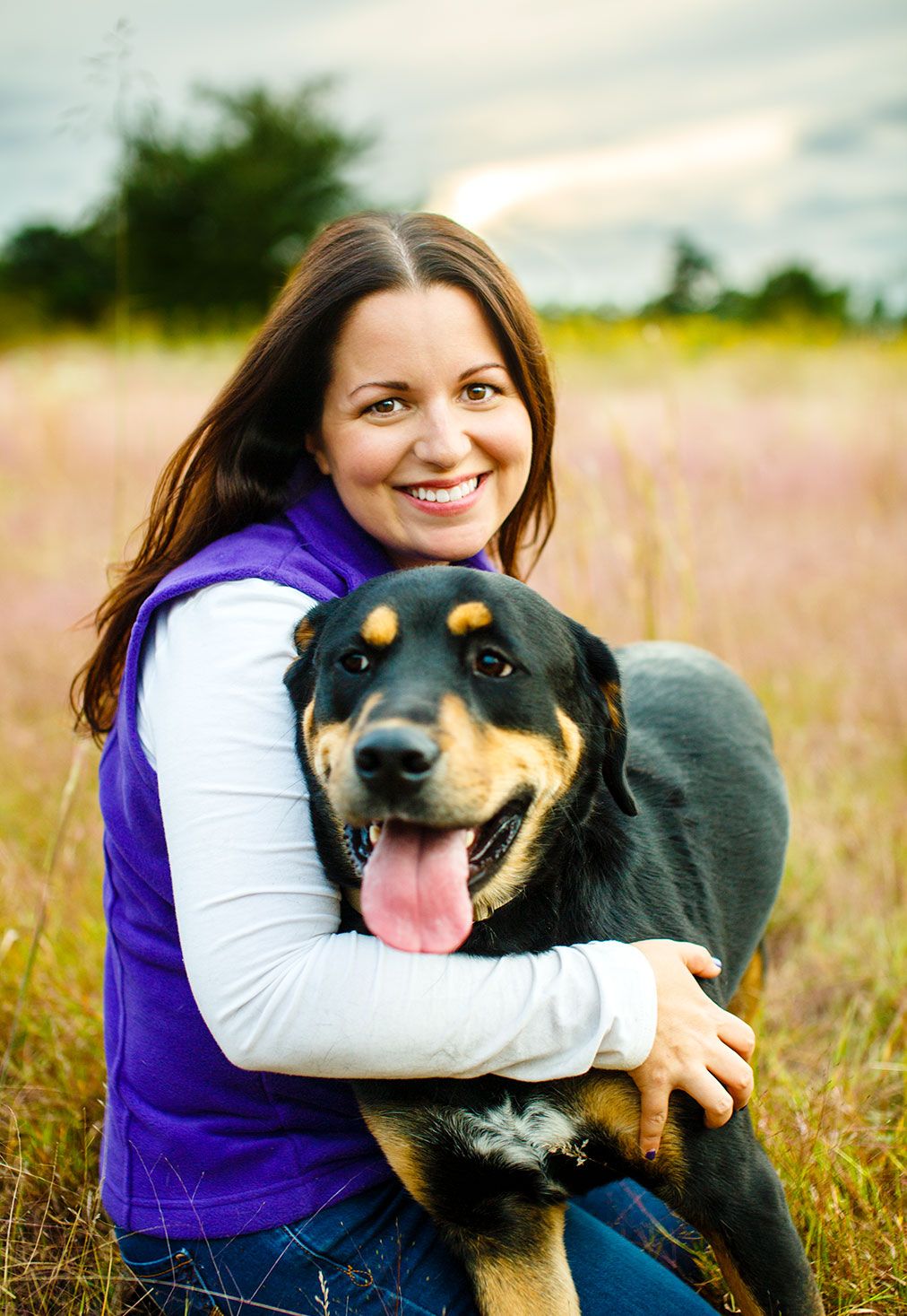 Smiling woman hugging a black and tan dog in a field.