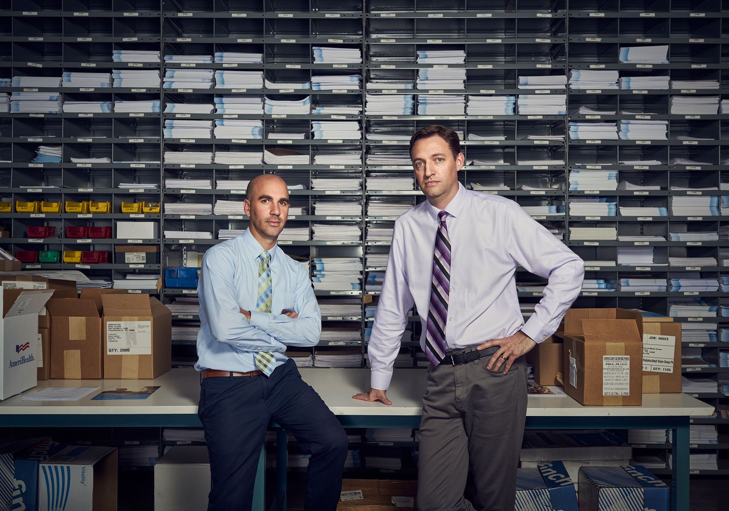 Two businessmen pose in storage room surrounded by organized filing systems and boxes.