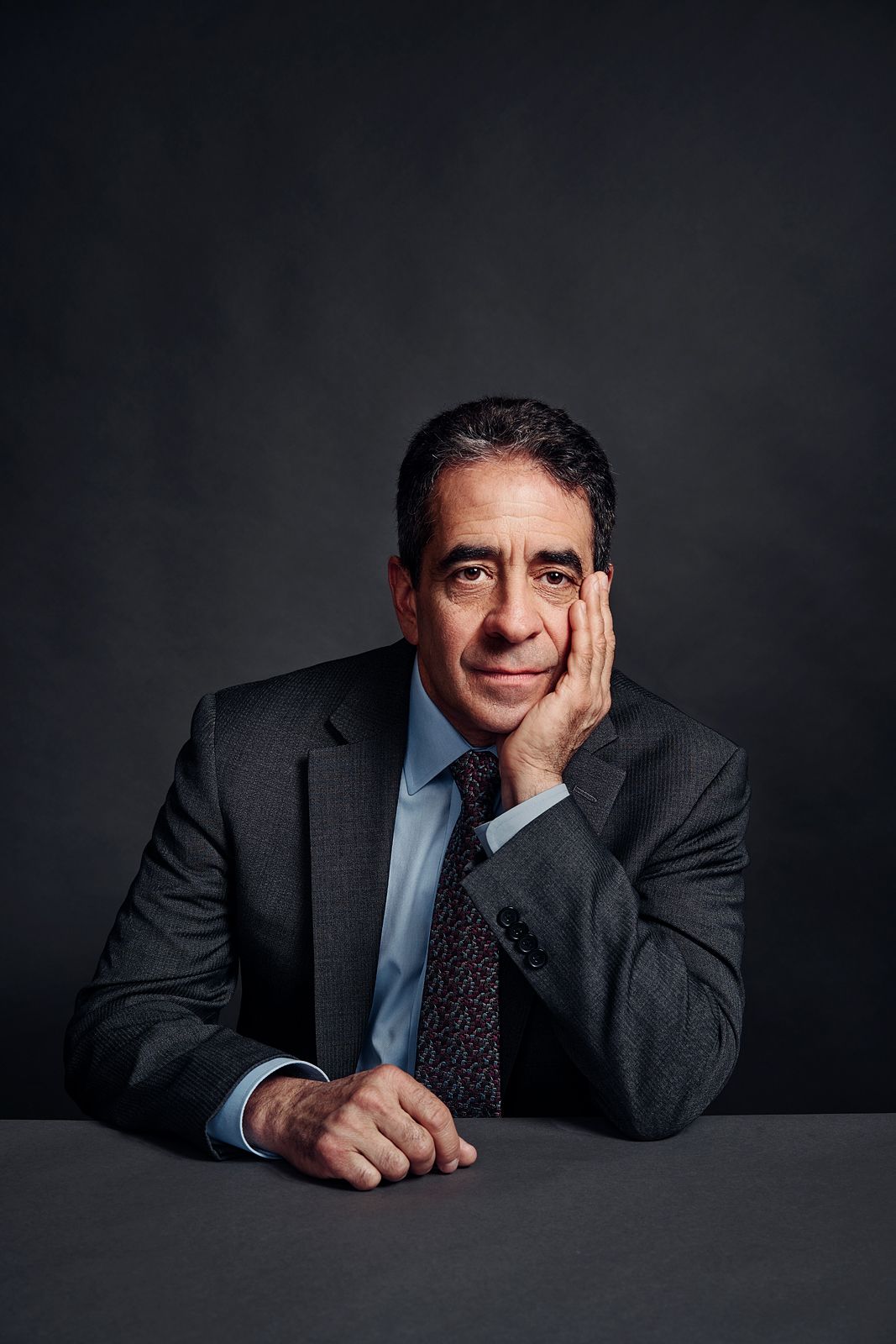 Professional man in dark suit and tie sits at table, resting cheek on hand, against dark background.