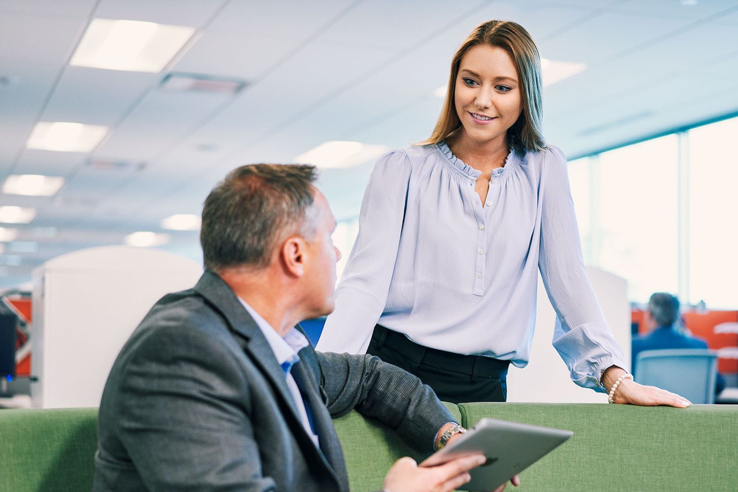 A smiling businesswoman in a blue blouse discusses work with a male colleague holding a tablet in a modern office.