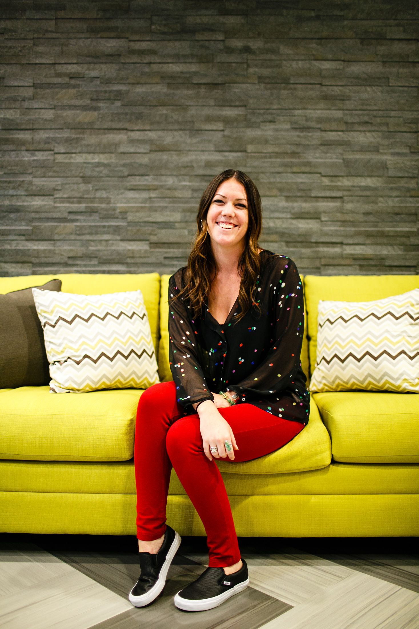 Smiling woman in red pants and black top sitting on a yellow couch with chevron pillows.