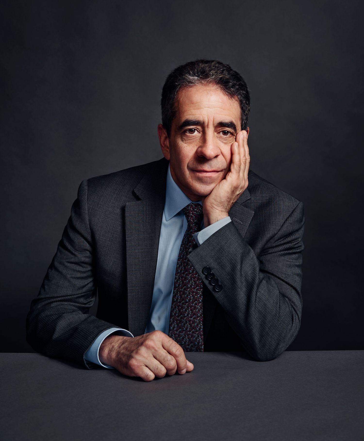 Professional man in dark suit and tie rests chin on hand at table for formal portrait.