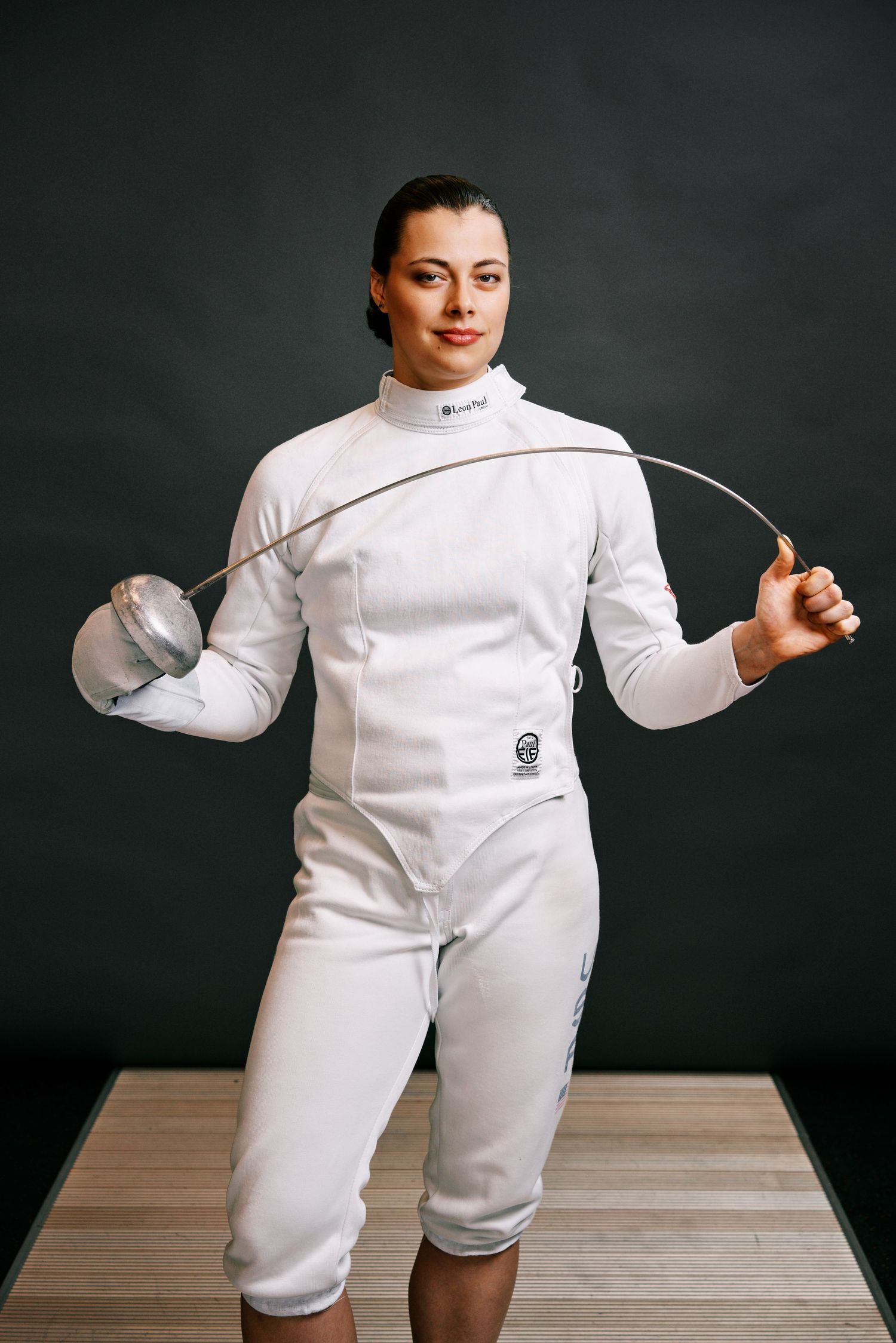 Female fencer in white protective gear holds épée sword and mask, posing confidently against dark background.