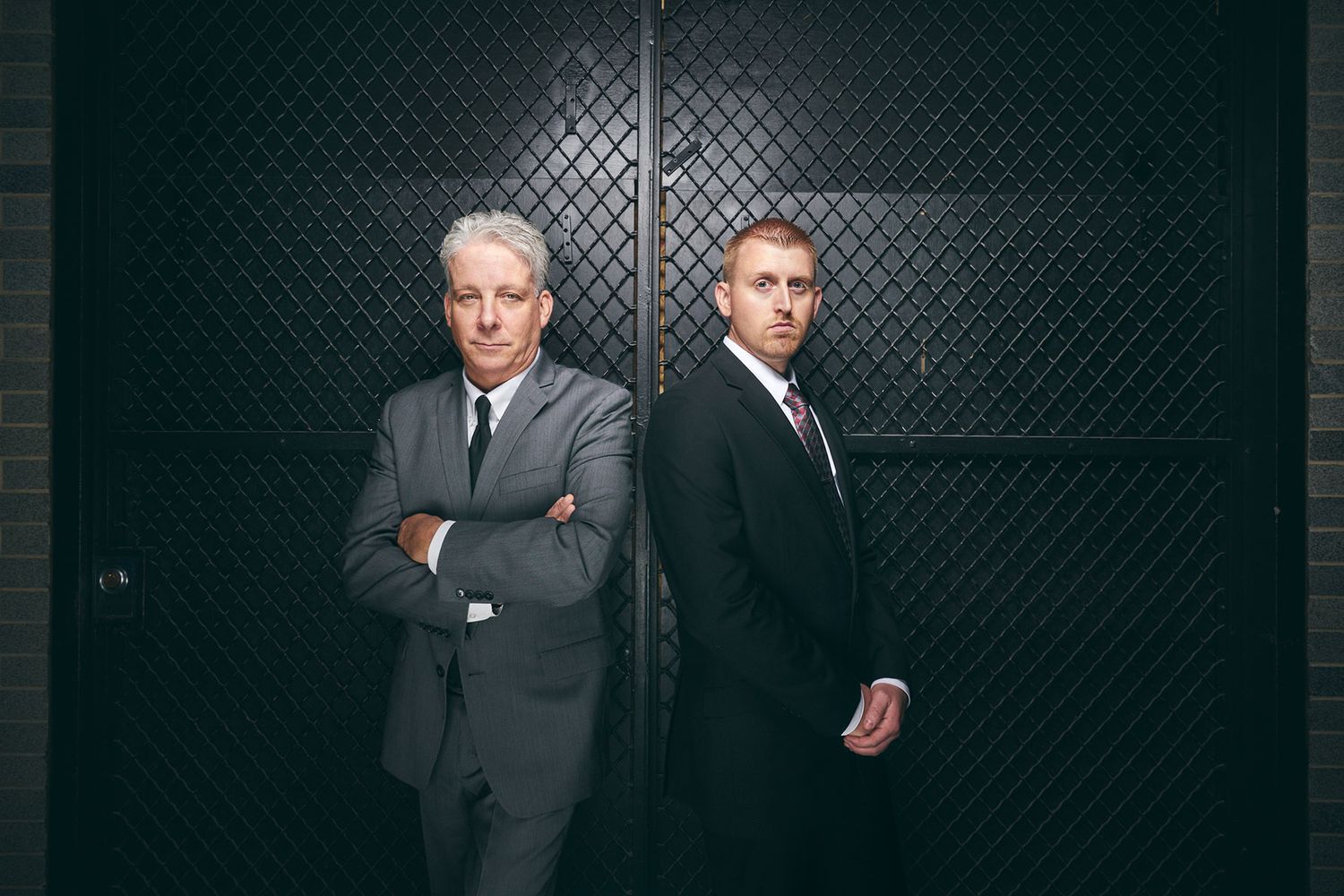 Two serious businessmen in suits posing back-to-back against dark metal door.