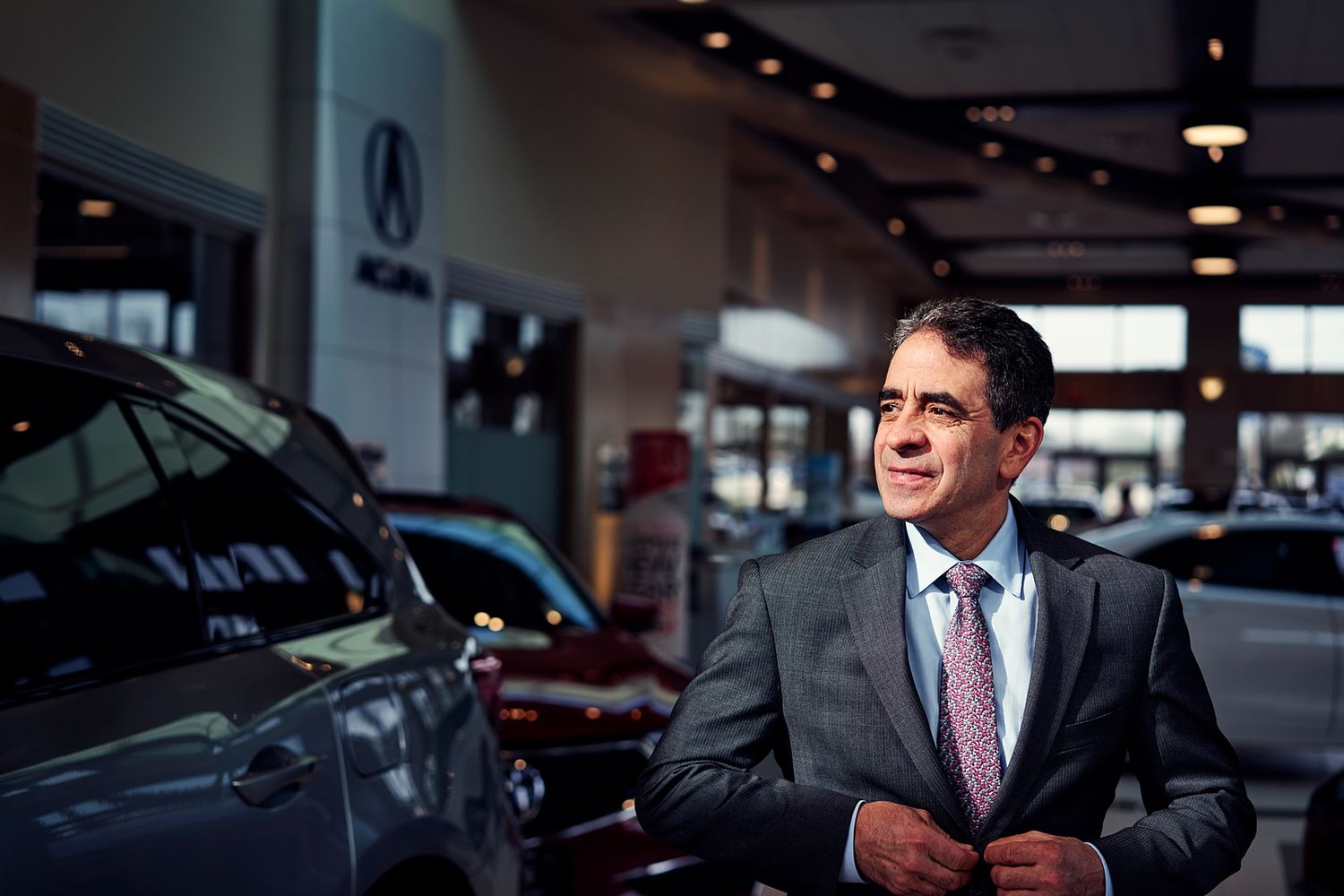 Professional man in suit stands confidently among luxury Acura vehicles inside a modern car dealership showroom.