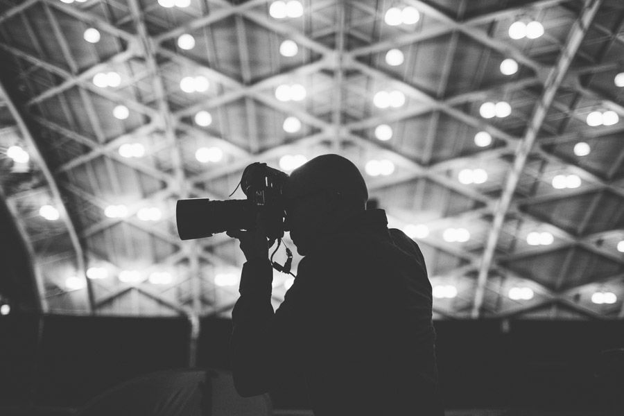 Silhouette of a photographer holding a camera against a bright ceiling of lights in a black and white photo.