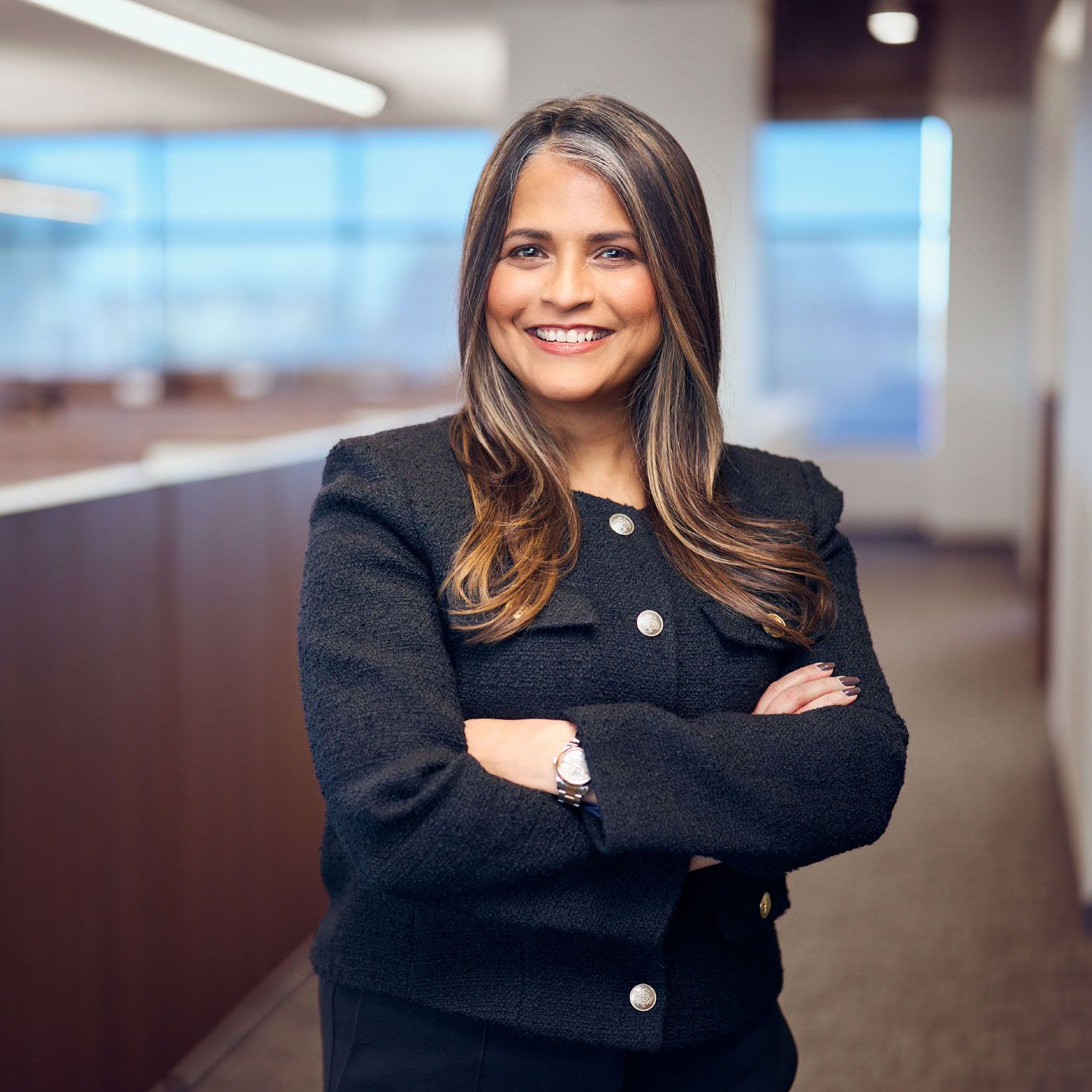 Professional businesswoman in dark blazer smiling with arms crossed in modern office setting.