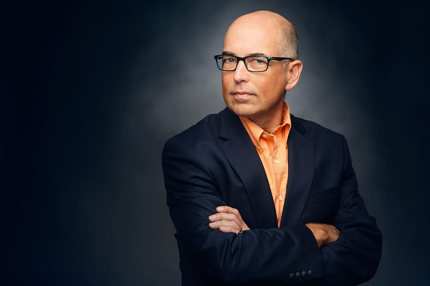 Professional man in orange shirt and dark blazer with arms crossed, wearing glasses against gray background.