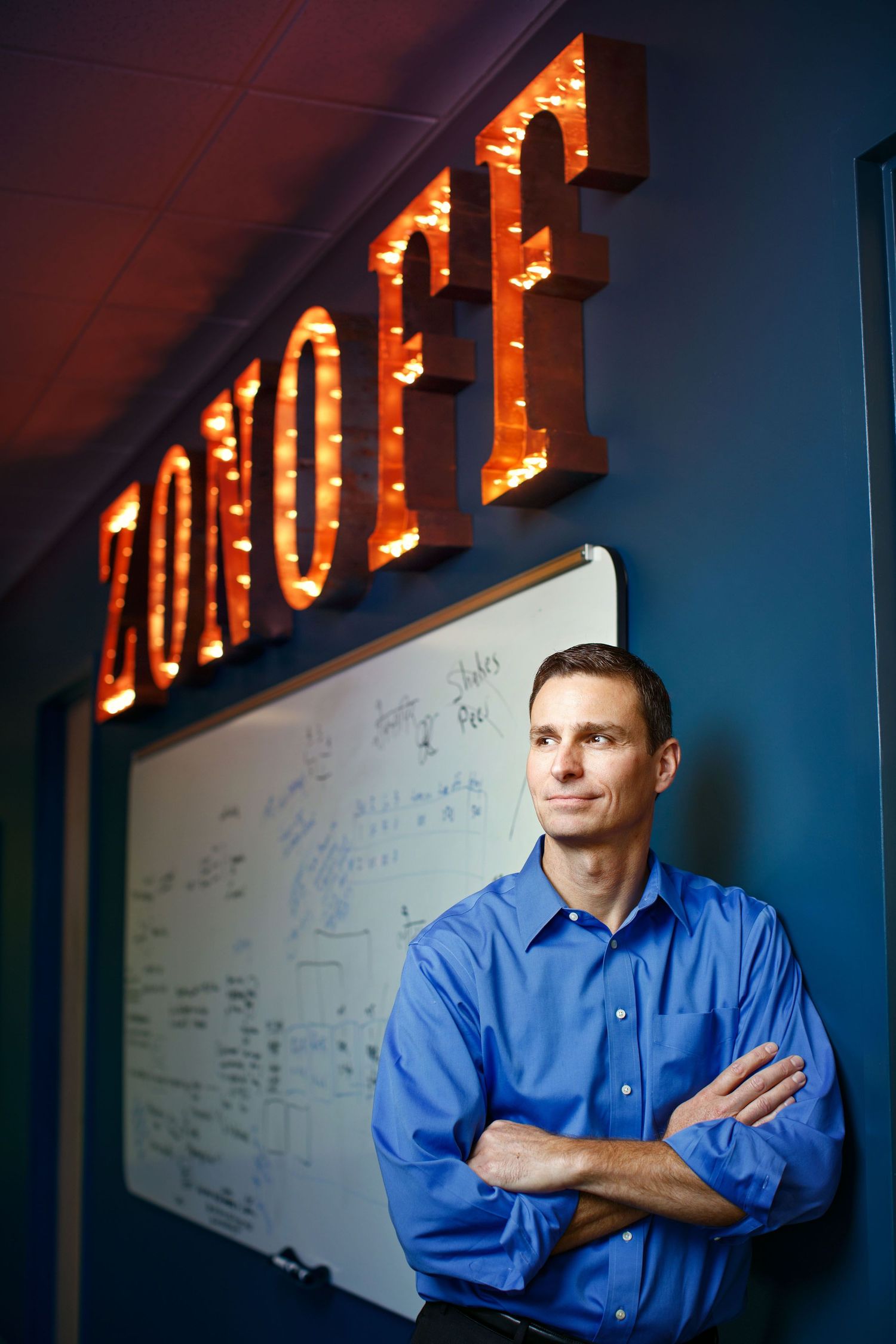 Professional man in blue shirt stands arms crossed under illuminated ZONOFF sign near whiteboard in office.