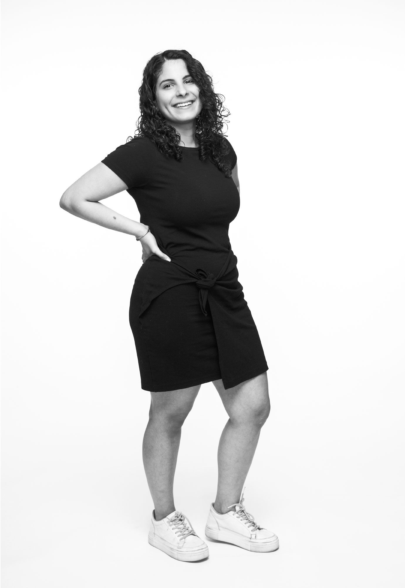 Black and white photo of curly-haired woman in black dress posing with hand on hip.