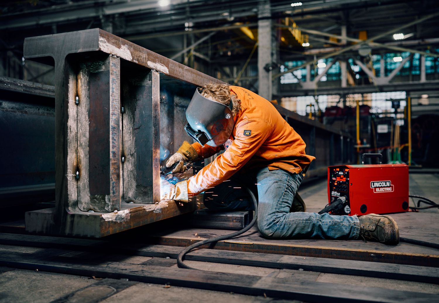 Industrial welder in orange safety gear working with metal beams in a factory warehouse.