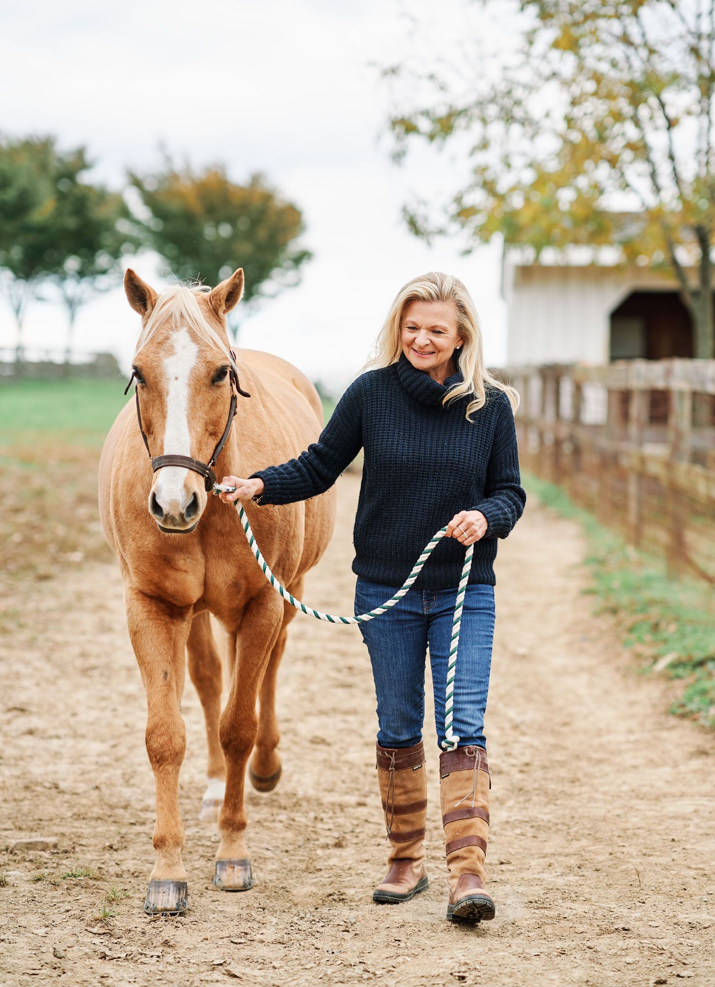 Woman in navy sweater and brown riding boots walks a chestnut horse along a farm path.