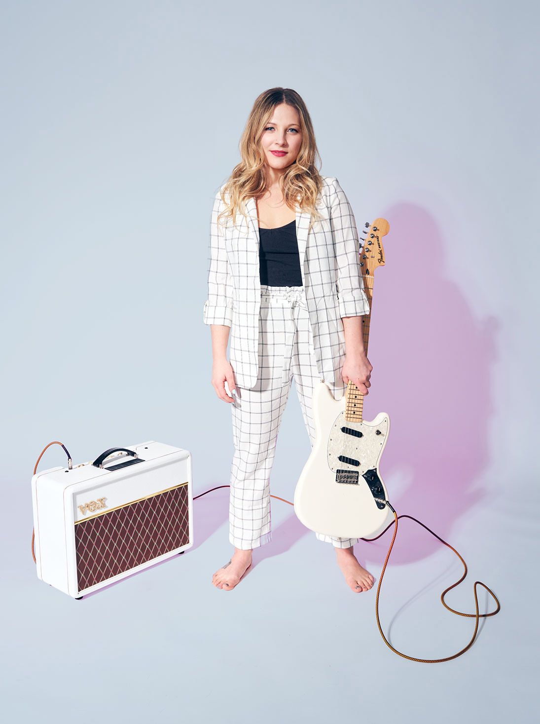 Barefoot woman in white grid suit holding electric guitar beside a Vox amp.
