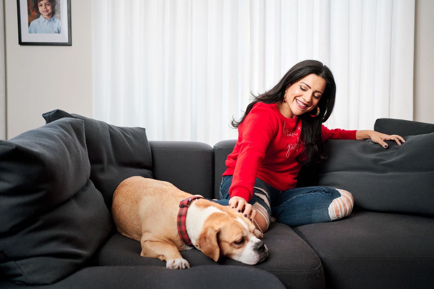 Woman in red sweater smiling while petting a beagle dog on a dark gray couch indoors.