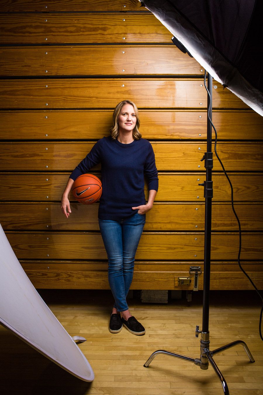Woman in navy sweater and jeans holding basketball against wooden gym wall with studio lighting equipment.