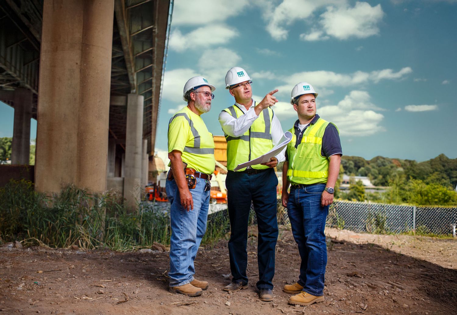 Three construction workers in hard hats reviewing plans under a bridge at a job site.