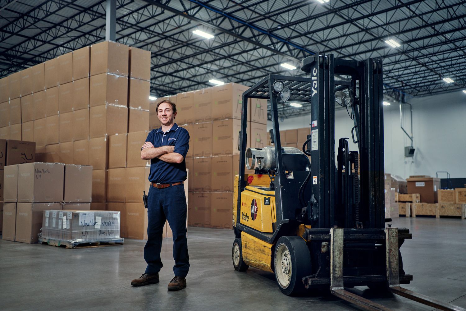 Smiling warehouse worker standing beside a yellow forklift surrounded by stacked cardboard boxes.