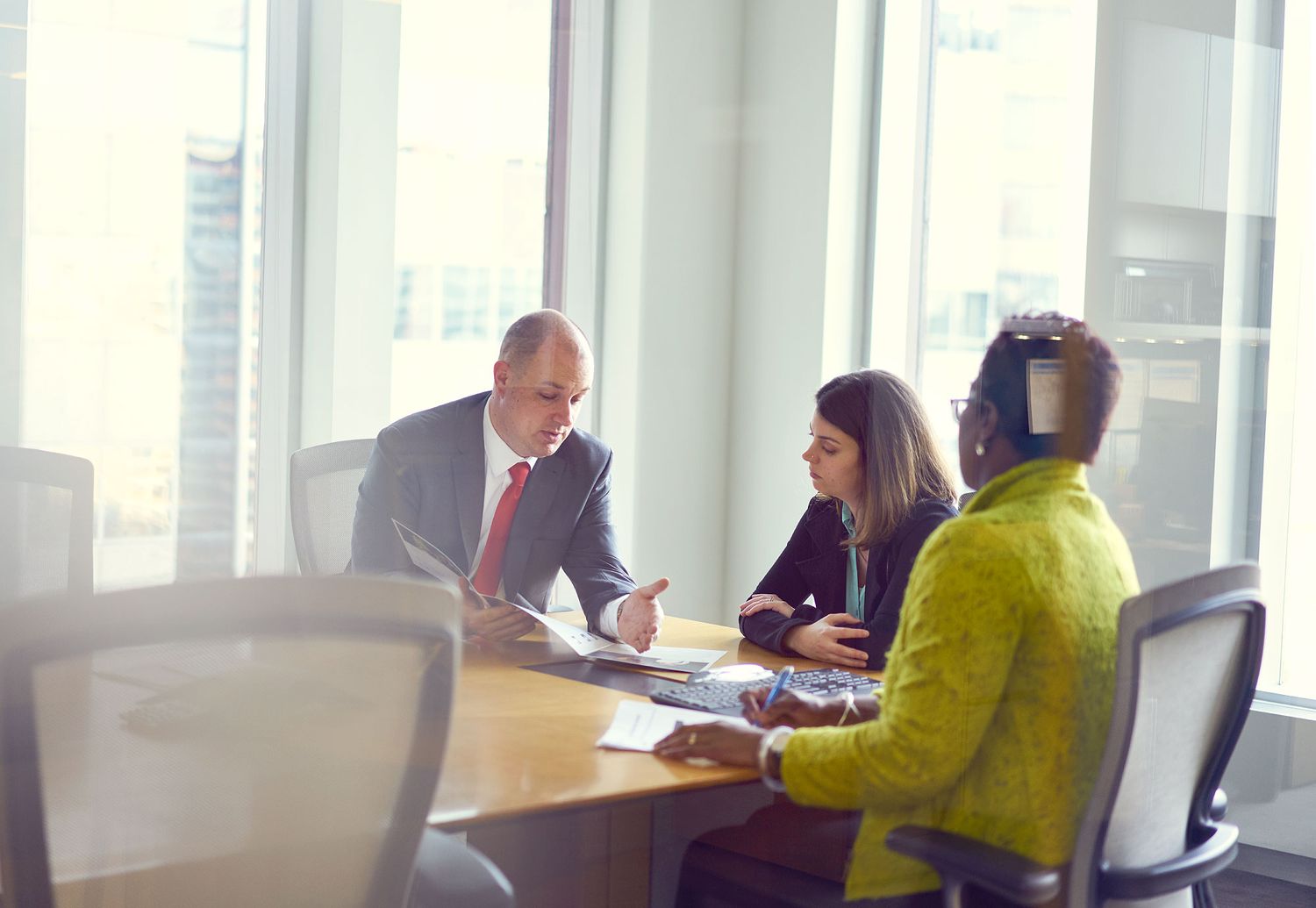 Three business professionals reviewing documents during a formal meeting in a glass-walled office.