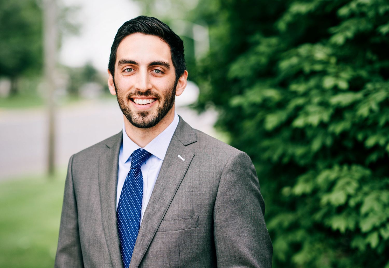 Young professional man in gray suit and blue tie smiling outdoors with green trees behind him.