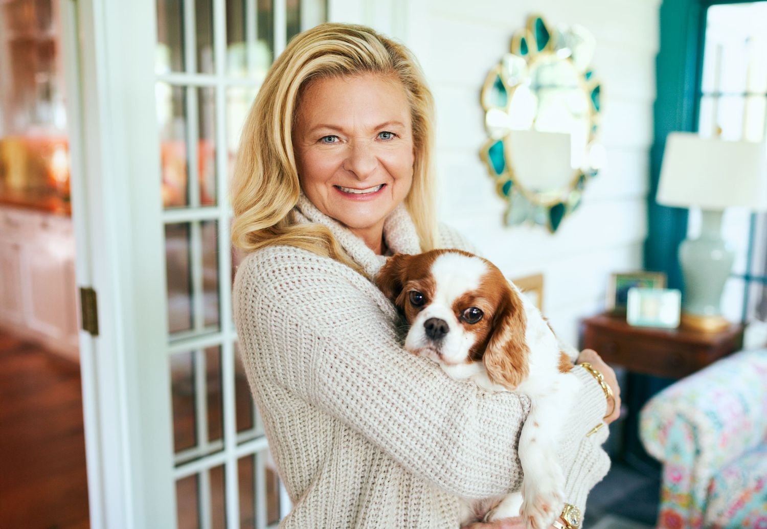 Smiling blonde woman holds a Cavalier King Charles Spaniel dog indoors.