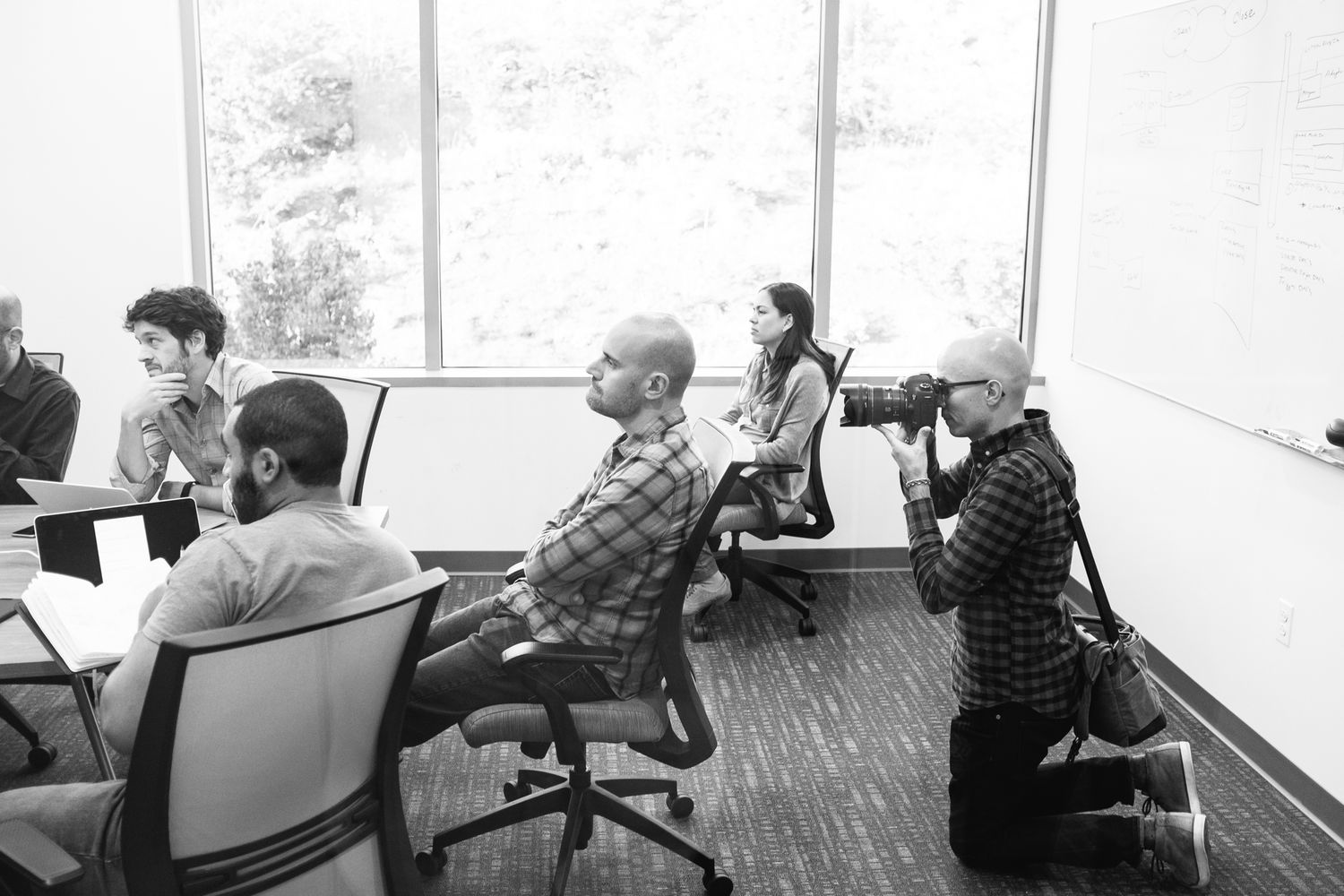 People in a modern office conference room, with a photographer kneeling to capture colleagues seated during a meeting.
