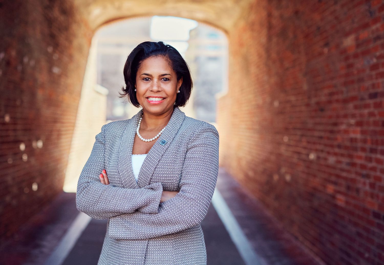 Professional woman in gray blazer with arms crossed smiling in brick archway outdoor setting.